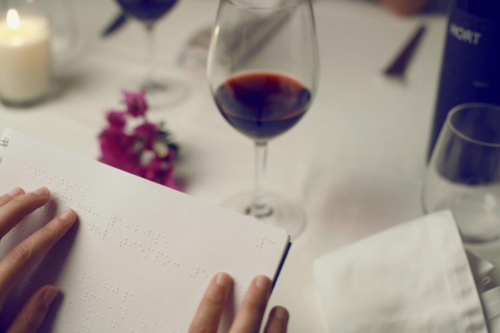 Braille menue in foreground. In the background a glass with red wine on a white tablecloth